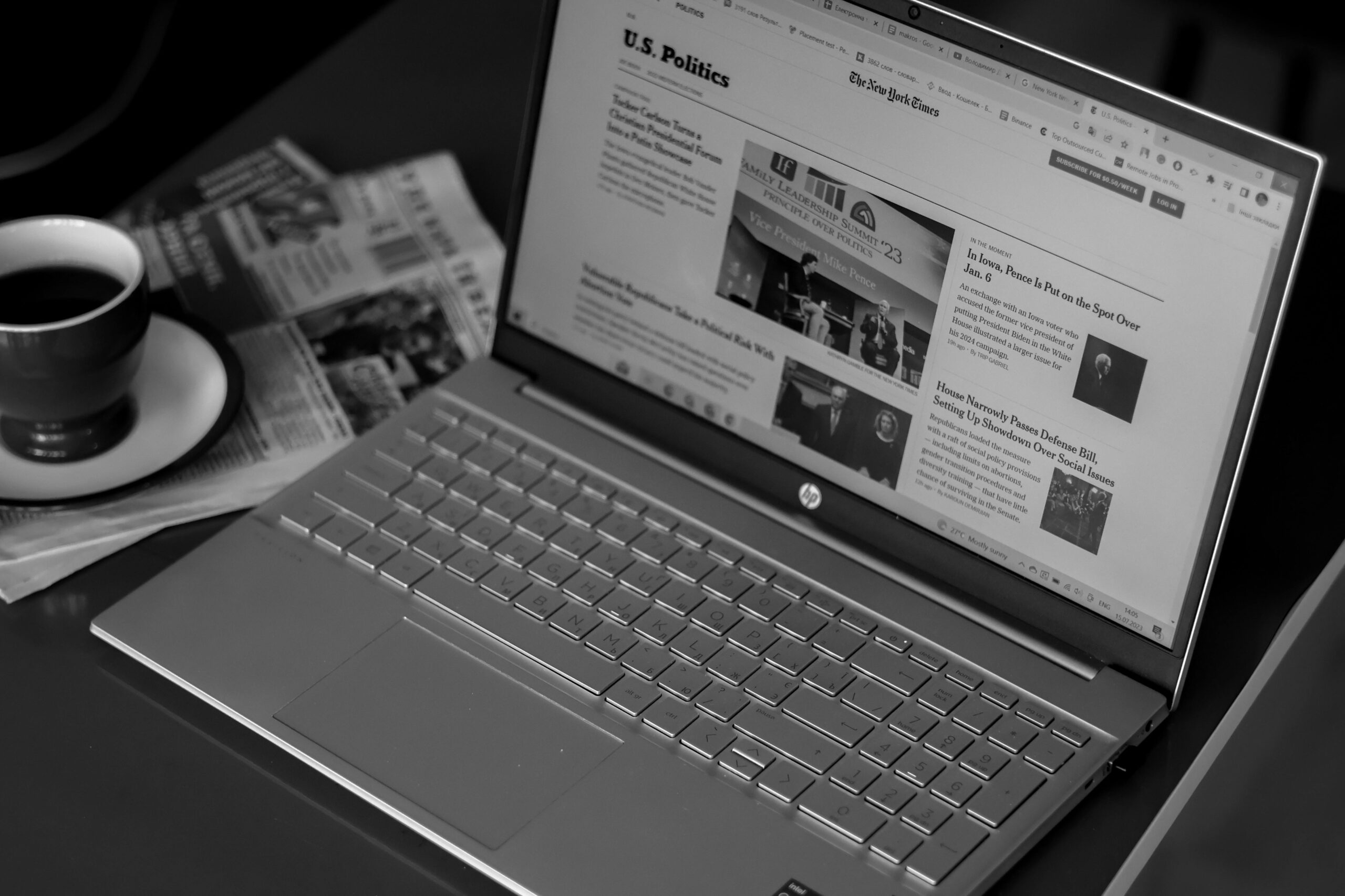 A monochrome image of an HP laptop open on a desk, with a cup of coffee and newspapers in the background, evoking a productive and informed atmosphere.